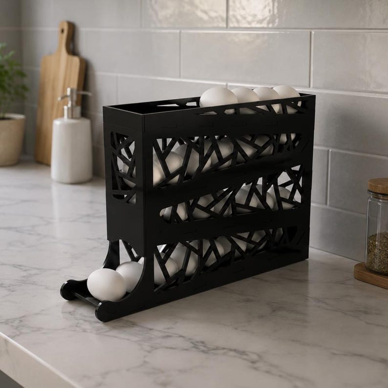 May include: A black, geometric egg holder filled with white eggs. The holder has a tiered design with a rolling mechanism at the bottom. The holder is made of a dark material and sits on a marble countertop.