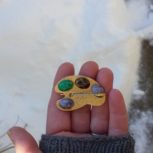 May include: Gold-toned artist's palette brooch with three colorful stones: green, brown, and purple.