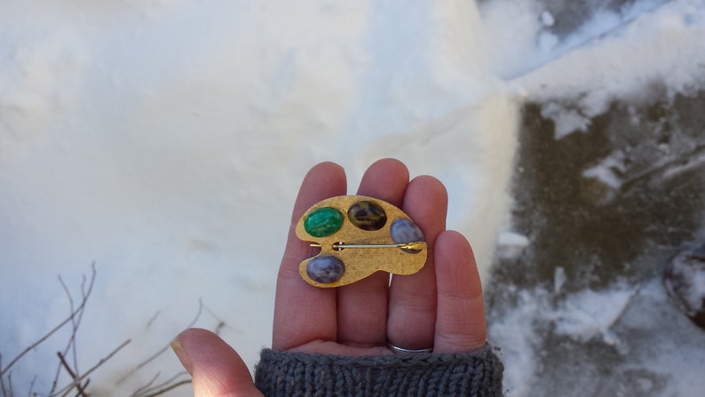 May include: Gold-toned artist's palette brooch with three colorful stones: green, brown, and purple.
