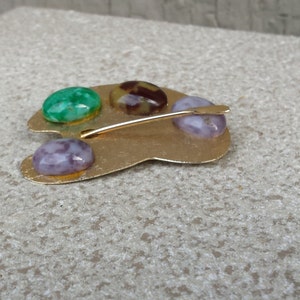 May include: Gold-toned metal artist's palette brooch with four colorful stones. The stones are green, purple, brown, and white.