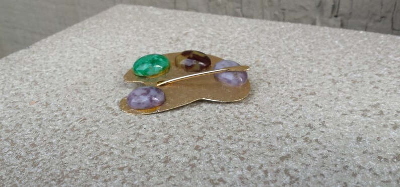 May include: Gold-toned metal artist's palette brooch with four colorful stones. The stones are green, purple, brown, and white.
