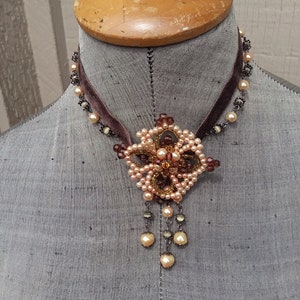 May include: A brown velvet choker necklace with a delicate beaded flower pendant. The flower is made of small, light pink and brown beads, and the pendant hangs from a chain of small, silver beads. The necklace is displayed on a mannequin.
