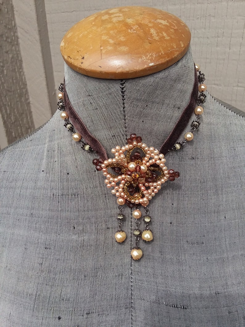 May include: A brown velvet choker necklace with a delicate beaded flower pendant. The flower is made of small, light pink and brown beads, and the pendant hangs from a chain of small, silver beads. The necklace is displayed on a mannequin.