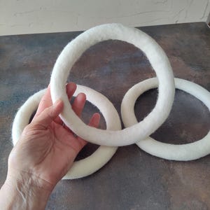 May include: Three white felt rings, one held by a hand, rest on a dark surface. The rings are thick and have a soft, textured appearance. The background is a muted, neutral color.