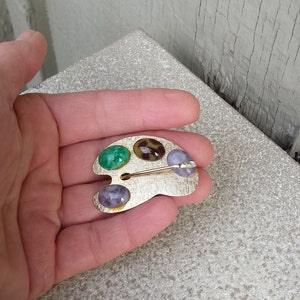 May include: A gold-toned artist's palette brooch with three colorful stones. The stones are green, brown with a leopard print, and purple.