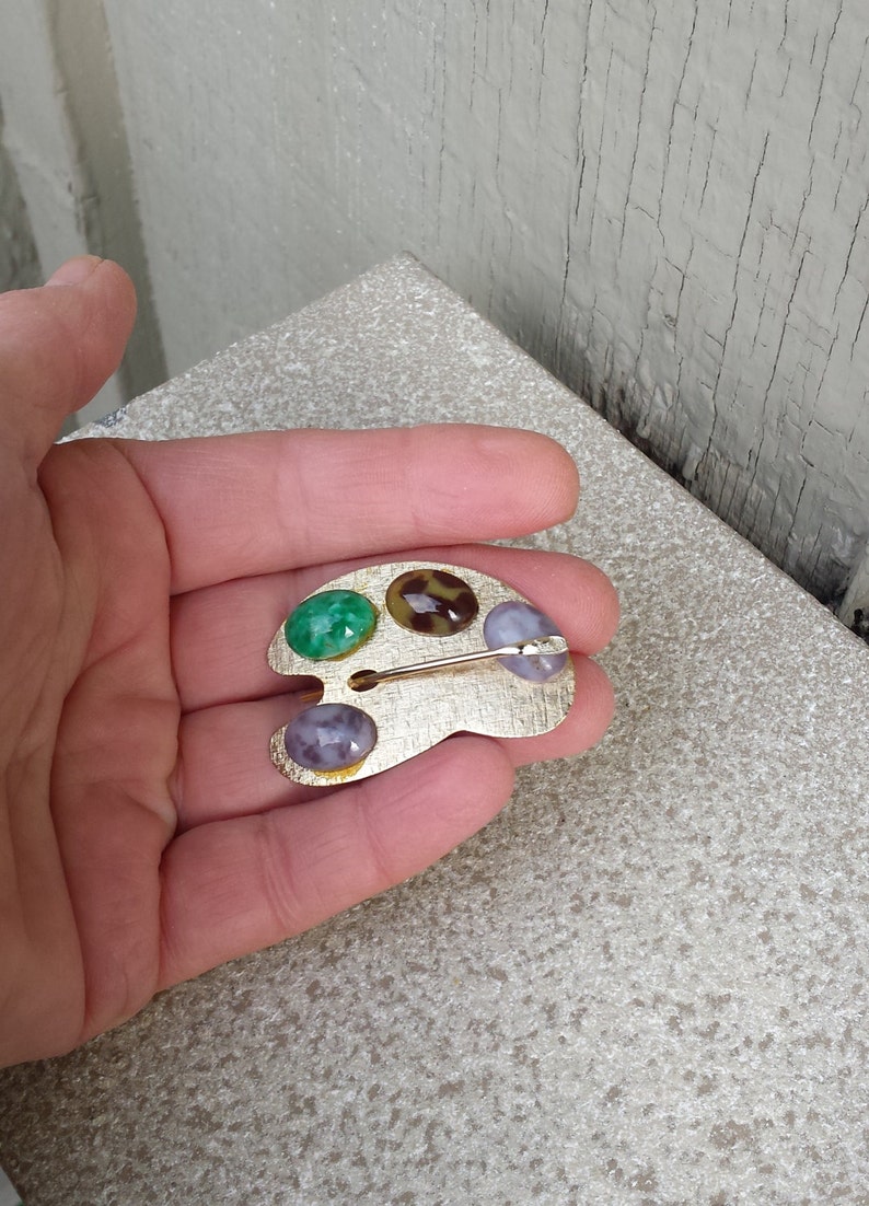 May include: A gold-toned artist's palette brooch with three colorful stones. The stones are green, brown with a leopard print, and purple.