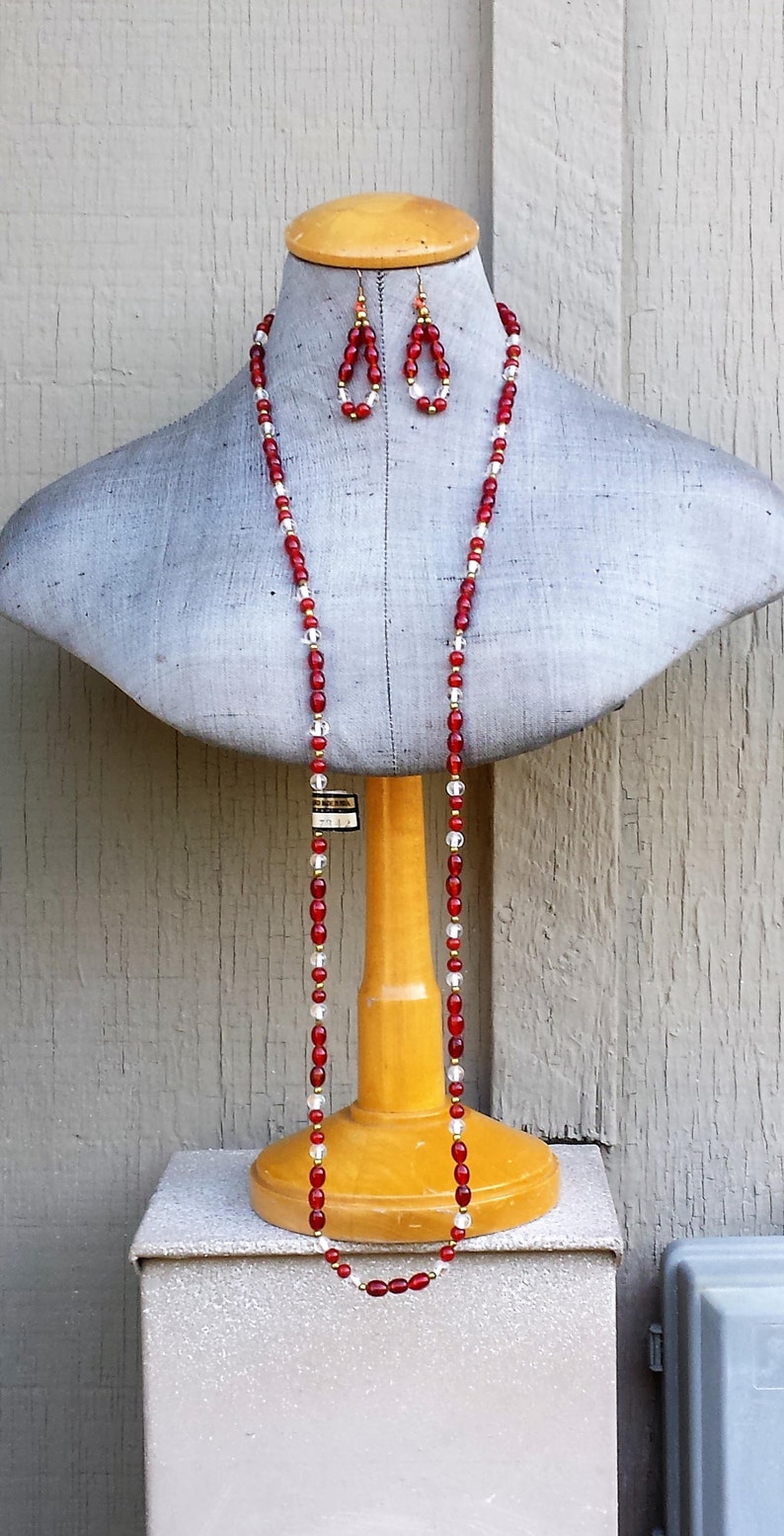 May include: A red and clear glass beaded necklace and matching earrings on a mannequin. The necklace is long and hangs down to the mannequin's waist. The earrings are small and dangle.