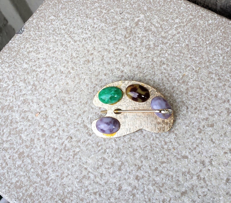 May include: A gold-toned metal artist's palette brooch with four oval-shaped stones. The stones are green, brown with white spots, purple with white spots, and purple with white spots. The brooch has a pin on the back.