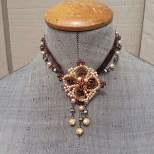 May include: A brown velvet choker necklace with a delicate beaded pendant featuring a floral design in shades of peach and brown.