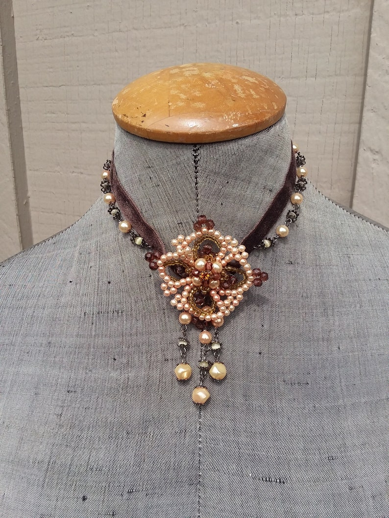 May include: A brown velvet choker necklace with a delicate beaded pendant featuring a floral design in shades of peach and brown.