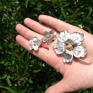 May include: A set of three silver and gold flower brooches. The largest brooch is a single flower with five petals. The two smaller brooches are each a single flower with four petals.