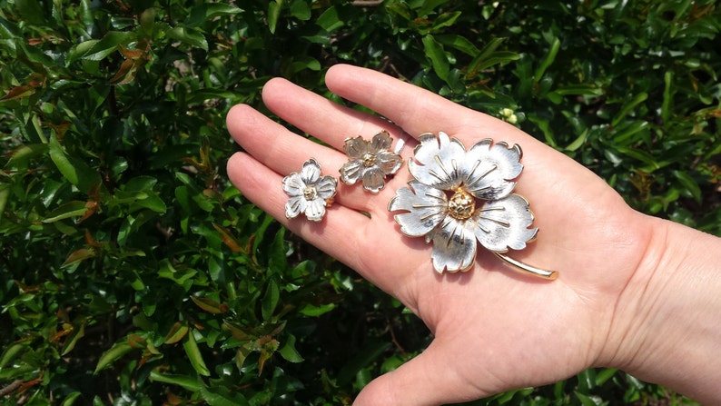 May include: A set of three silver and gold flower brooches. The largest brooch is a single flower with five petals. The two smaller brooches are each a single flower with four petals.