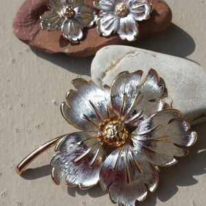 May include: A silver and gold flower brooch with a stem. The flower has five petals and a gold centre.