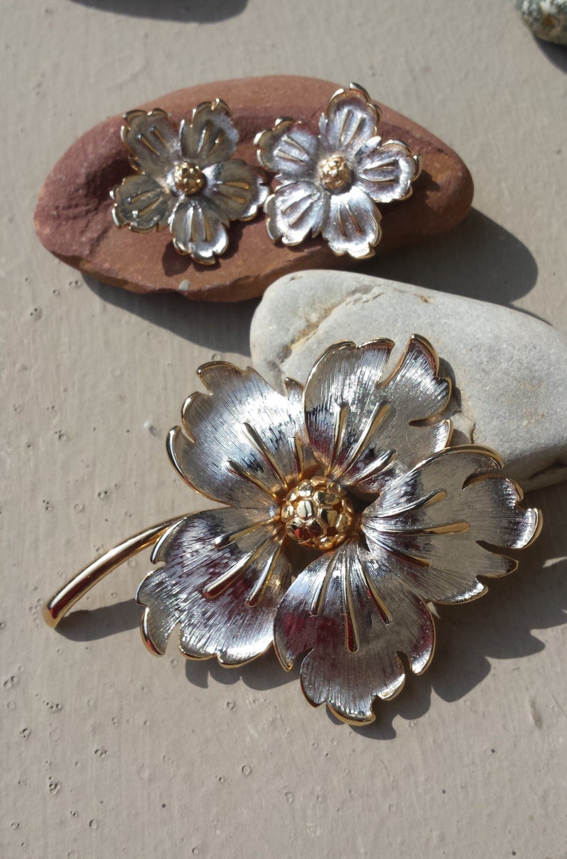 May include: A silver and gold flower brooch with a stem. The flower has five petals and a gold centre.