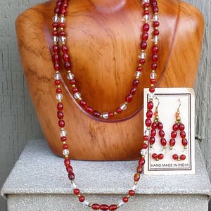 May include: A red and clear glass bead necklace and matching earrings. The necklace is double-stranded and features gold accents. The earrings are dangle style and have a similar design to the necklace. The jewelry is displayed on a wooden mannequin and a white box. The card reads "Hand Made in India".