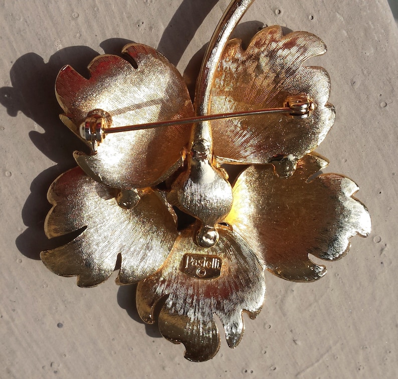 May include: Gold-toned flower brooch with a pin on the back. The brooch is marked "Pascucci" on the back.