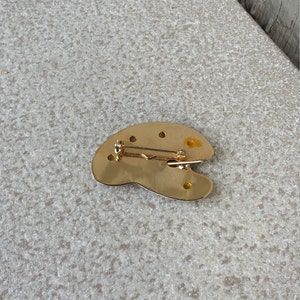 May include: Gold-toned metal artist's palette brooch with a pin on the back.
