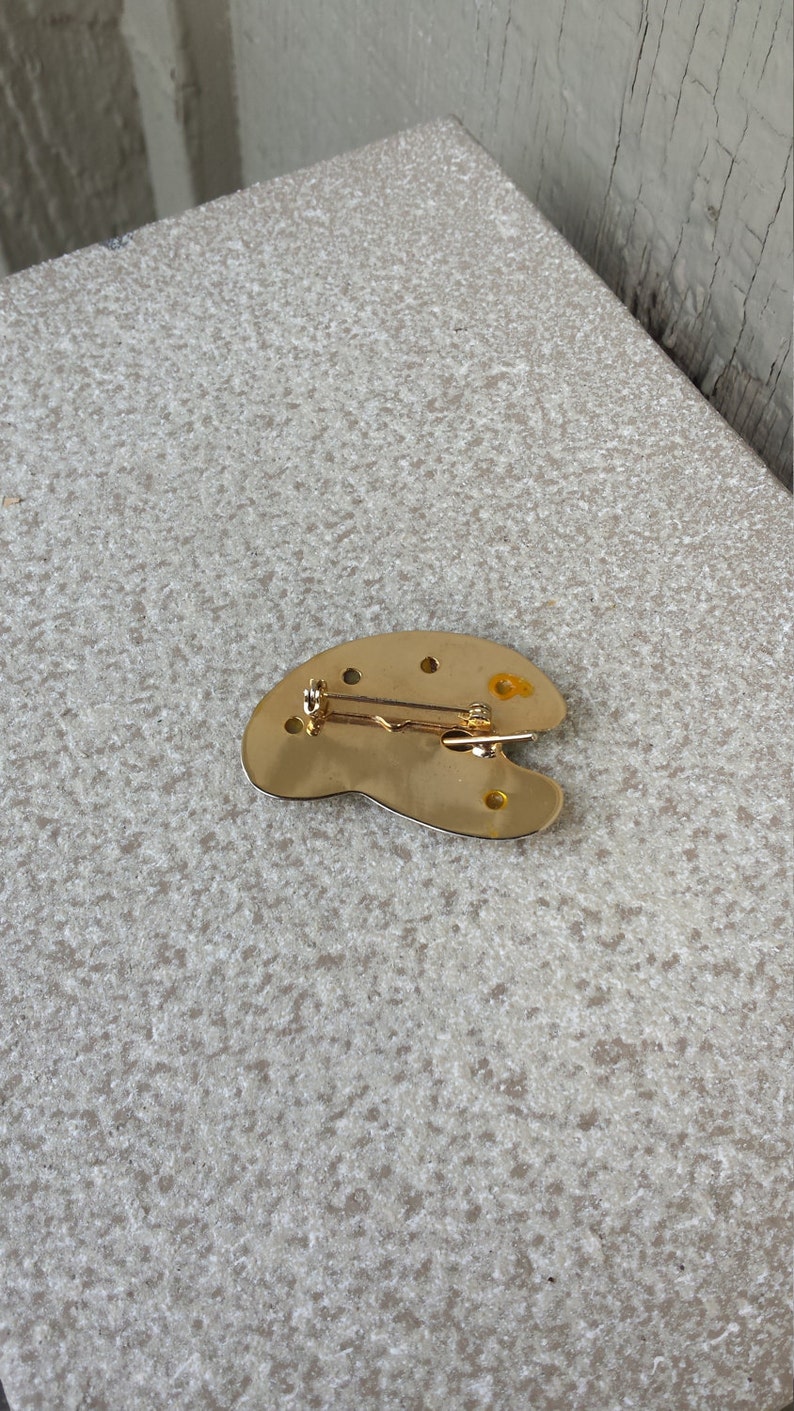 May include: Gold-toned metal artist's palette brooch with a pin on the back.