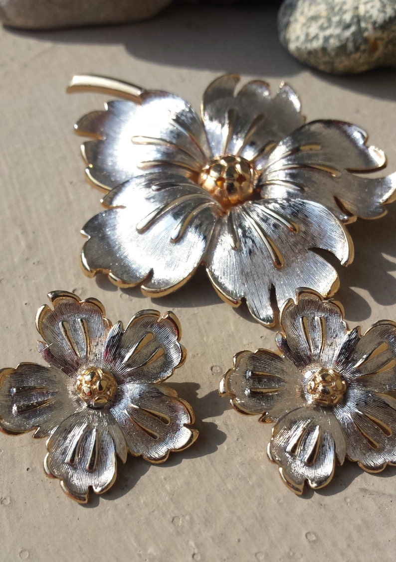 May include: A set of three silver and gold tone flower brooches. The largest brooch is a single flower with five petals. The two smaller brooches are each a single flower with five petals.