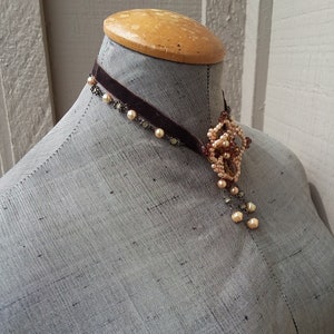 May include: A brown velvet choker necklace with a delicate pendant featuring a cluster of small, cream-colored pearls and beads.