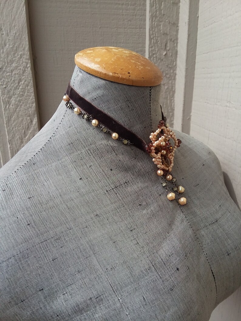 May include: A brown velvet choker necklace with a delicate pendant featuring a cluster of small, cream-colored pearls and beads.
