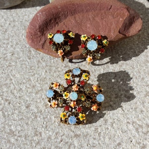 May include: A set of vintage gold tone earrings and a brooch with a floral design. The jewelry features pastel blue, yellow, red, and green enamel flowers with clear rhinestones.