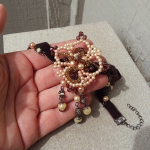 May include: A brown velvet choker necklace with a large beaded flower pendant. The flower is made of light brown and dark brown beads and has a crystal center. The necklace has a silver clasp.