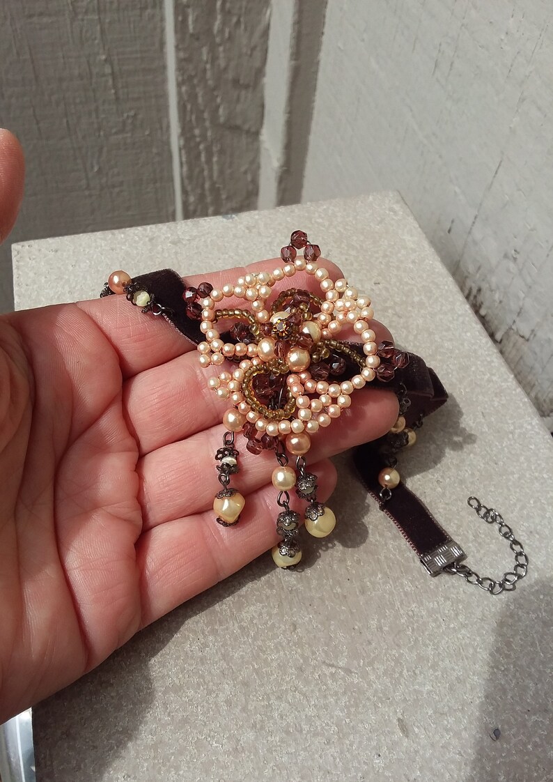 May include: A brown velvet choker necklace with a large beaded flower pendant. The flower is made of light brown and dark brown beads and has a crystal center. The necklace has a silver clasp.