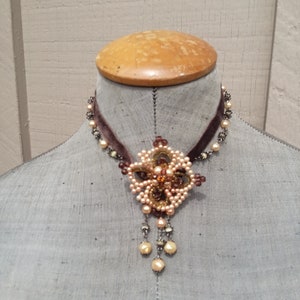 May include: A brown velvet choker necklace with a delicate floral pendant made of pearls and beads. The pendant features a central brown stone and hangs from a chain with pearl accents.
