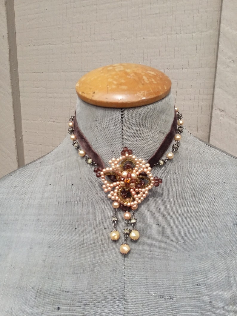 May include: A brown velvet choker necklace with a delicate floral pendant made of pearls and beads. The pendant features a central brown stone and hangs from a chain with pearl accents.