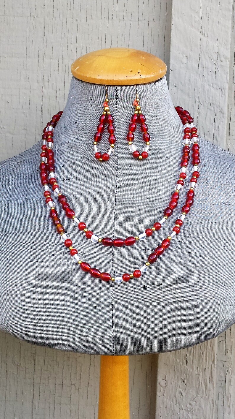 May include: A two-strand necklace and matching earrings with red and clear glass beads. The necklace has a gold accent between the beads.