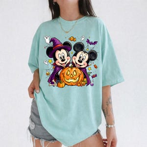 May include: A light mint green t-shirt featuring a graphic print of two cartoon mice dressed in Halloween costumes, one wearing a purple witch hat and cape, both standing behind a large orange jack-o'-lantern surrounded by candy and ghosts.