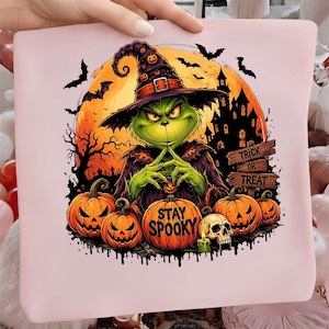 Puede incluir: Un cuadrado de tela rosa claro con un gráfico de una criatura verde con sombrero de bruja, frente a una gran luna naranja, un castillo oscuro y murciélagos. El diseño incluye calabazas talladas, una calavera y letreros que dicen TRICK OR TREAT y STAY SPOOKY.