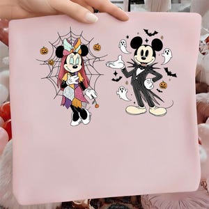 Peut inclure: Un article en tissu rose clair de forme carrée avec une impression graphique de deux personnages de dessin animé. L'un porte une robe en patchwork devant une toile d'araignée, l'autre un costume rayé entouré de fantômes, chauves-souris et citrouilles.