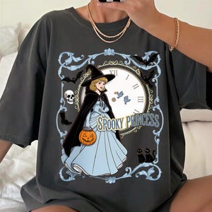 May include: A charcoal gray t-shirt featuring a graphic print of a blonde character in a light blue gown and black witch hat, holding a jack-o'-lantern. The design includes a clock face, bats, a skull, two black cats, and the text SPOOKY PRINCESS.