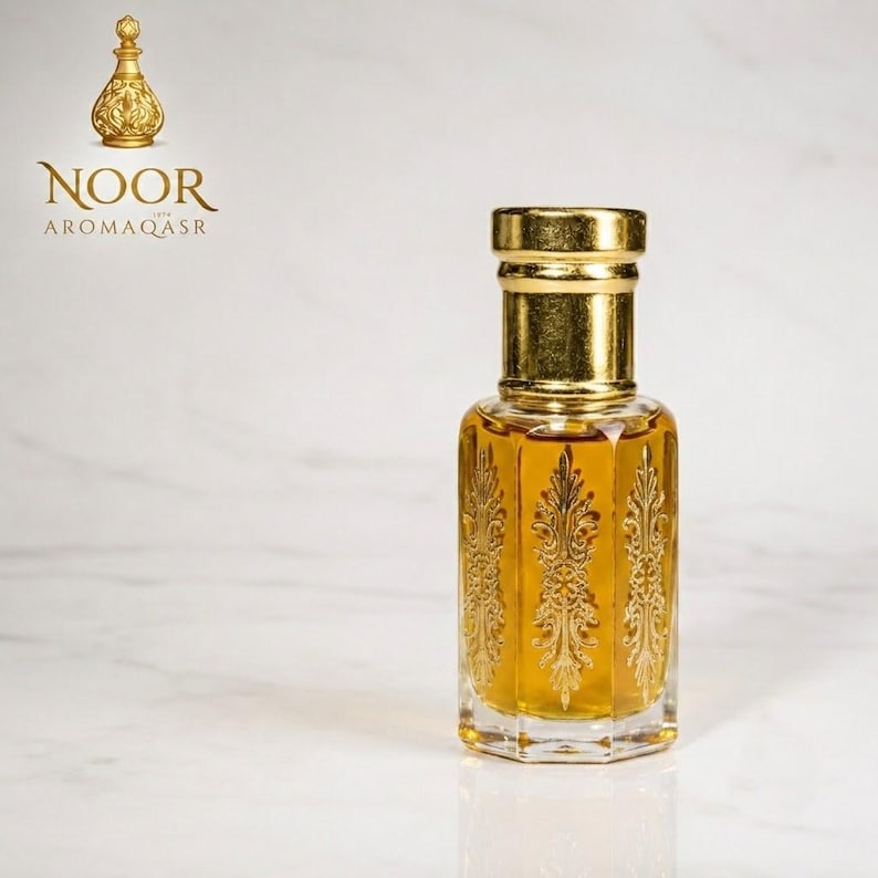 May include: A clear glass perfume bottle with a hexagonal shape and gold-colored cap. The bottle features intricate gold filigree patterns on each side and contains a golden liquid. The text NOOR AROMAQASR appears in the upper left corner.