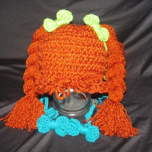 May include: A crocheted hat with long orange braids and green bows. The hat is made of a soft, textured yarn and has a loose, comfortable fit.