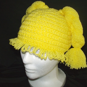 May include: A yellow crocheted hat with two long pigtails. The hat has a fringe around the bottom edge.