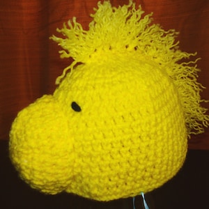 May include: A handmade, bright yellow crocheted hat shaped like a cartoon bird's head. The hat features a prominent beak, a small black eye, and a textured, fringed crest. The hat is designed to be worn on the head.