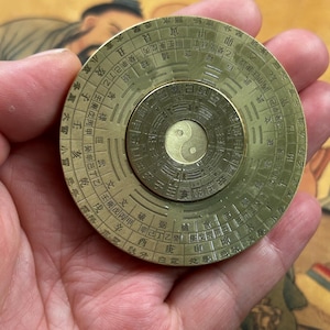 May include: A brass compass with intricate Chinese characters and a central Yin Yang symbol. The compass has concentric circles with detailed markings and is held in a hand.