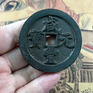 Może przedstawiać: Ciemnozielona, okrągła moneta z kwadratowym otworem pośrodku. Moneta posiada wytłoczone chińskie znaki. Moneta jest trzymana w dłoni. Powierzchnia monety ma lekko zużyty wygląd.