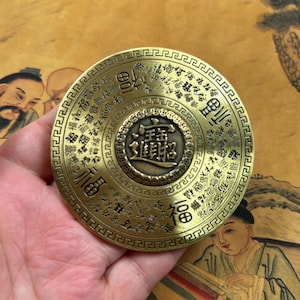 Puede incluir: Una moneda decorativa redonda de color latón antiguo con caracteres chinos. La moneda tiene un diseño central con más caracteres y está rodeada por un anillo de caracteres adicionales. La moneda se sostiene en una mano.