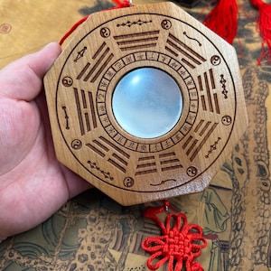 May include: Wooden octagonal Feng Shui mirror with engraved symbols and a central reflective surface. The mirror is adorned with red tassels and a decorative knot, suggesting a cultural or spiritual significance.