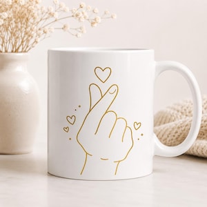 Könnte beinhalten: Ein weißer Keramikbecher mit einer goldenen Strichzeichnung einer Hand, die ein Fingerherz formt. Das Design enthält mehrere kleine goldene Herzen, die über und um die Hand herum schweben und ein schlichtes, klares Erscheinungsbild erzeugen.