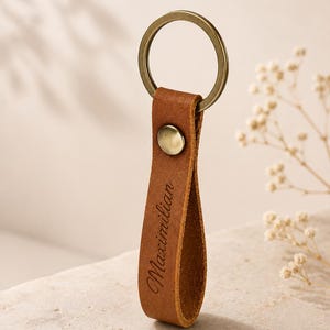 Könnte beinhalten: Ein hellbrauner Schlüsselanhänger aus Leder mit einem messingfarbenen Metallring und einem passenden Druckknopf. Das Lederband ist zu einer Schlaufe gefaltet und trägt den Namen Maximilian in einer dunklen Schreibschrift auf der Vorderseite.