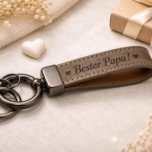 Könnte beinhalten: Ein brauner Leder-Schlüsselanhänger mit dem Text "Bester Papa!" und einem Herzsymbol. Der Schlüsselanhänger hat einen Metallverschluss und liegt neben einer Geschenkbox mit Schleife.
