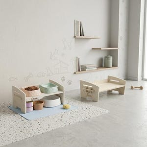 May include: A light wood pet feeding station with two tiers, holding food bowls and containers. A matching pet bed is nearby. The scene includes a blue mat, a bone-shaped toy, and wall art with pet-themed drawings.