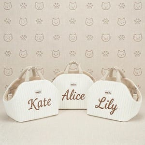 May include: Three white pet carriers with brown embroidered names: "Kate", "Alice", and "Lily". Each carrier has a cream-colored handle and interior. The background features a repeating pattern of cat faces and paw prints.