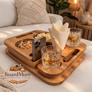 Puede incluir: Una bandeja de bambú con compartimentos para aperitivos, un teléfono y dos vasos. La bandeja contiene cacahuetes, palomitas de maíz, un teléfono, servilletas y dos vasos de líquido ámbar. La bandeja tiene el texto "BoardMum WOODEN BOARDS & TRAYS".