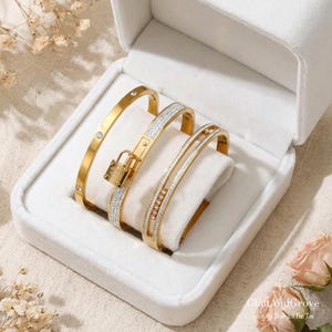 May include: A collection of gold-coloured bangles presented in a white box. The bangles include a plain band with three small crystals, a band with a lock, and a band with a row of crystals. The box is lined with white fabric, with a floral background.